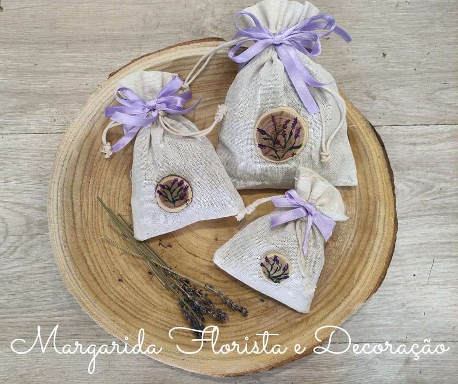 Sacos de tecido bege com laços lilás e flores, sobre prato de madeira redondo com ramo de lavanda