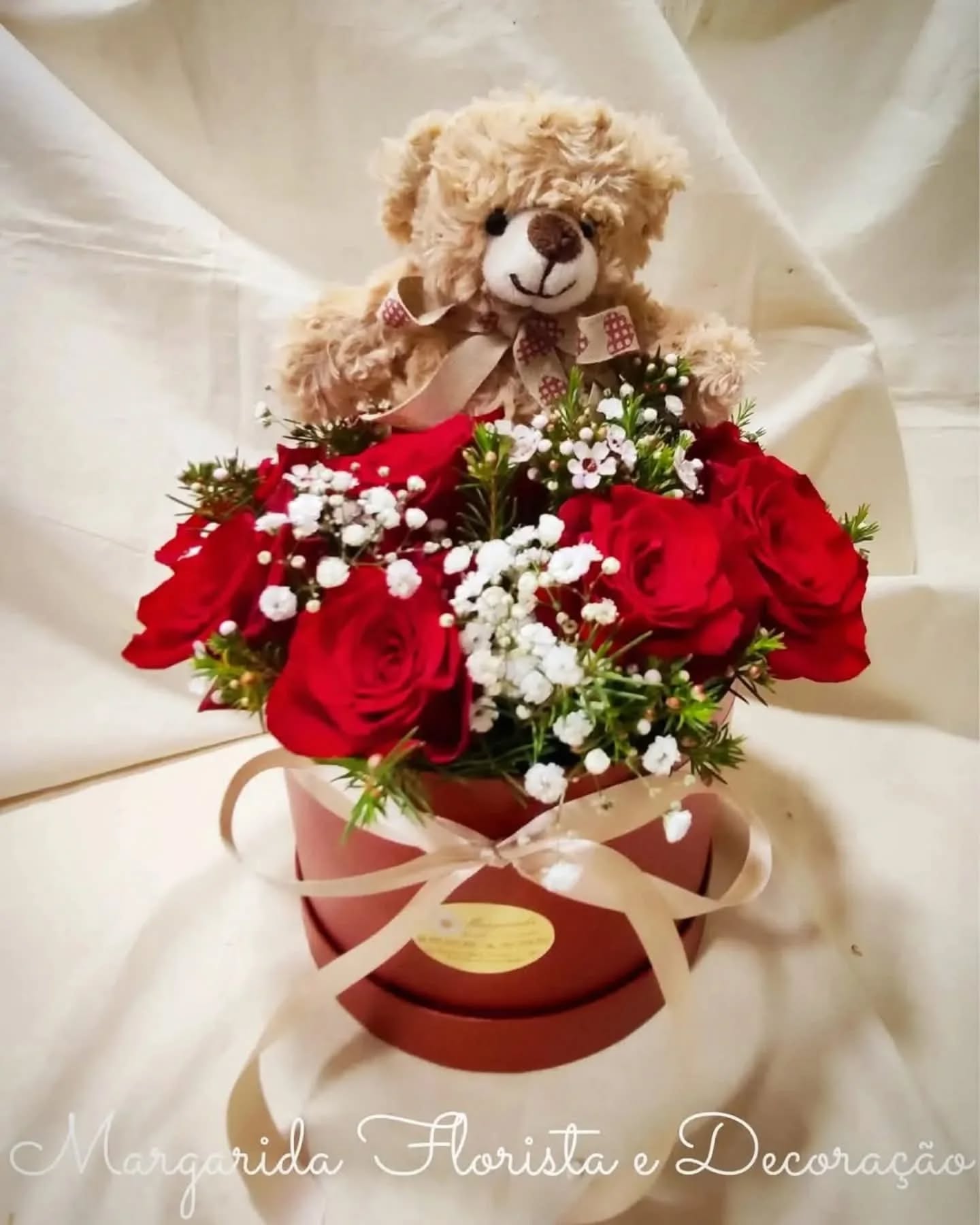 Arranjo floral com rosas vermelhas, flores brancas e ursinho de pelúcia numa caixa redonda.
