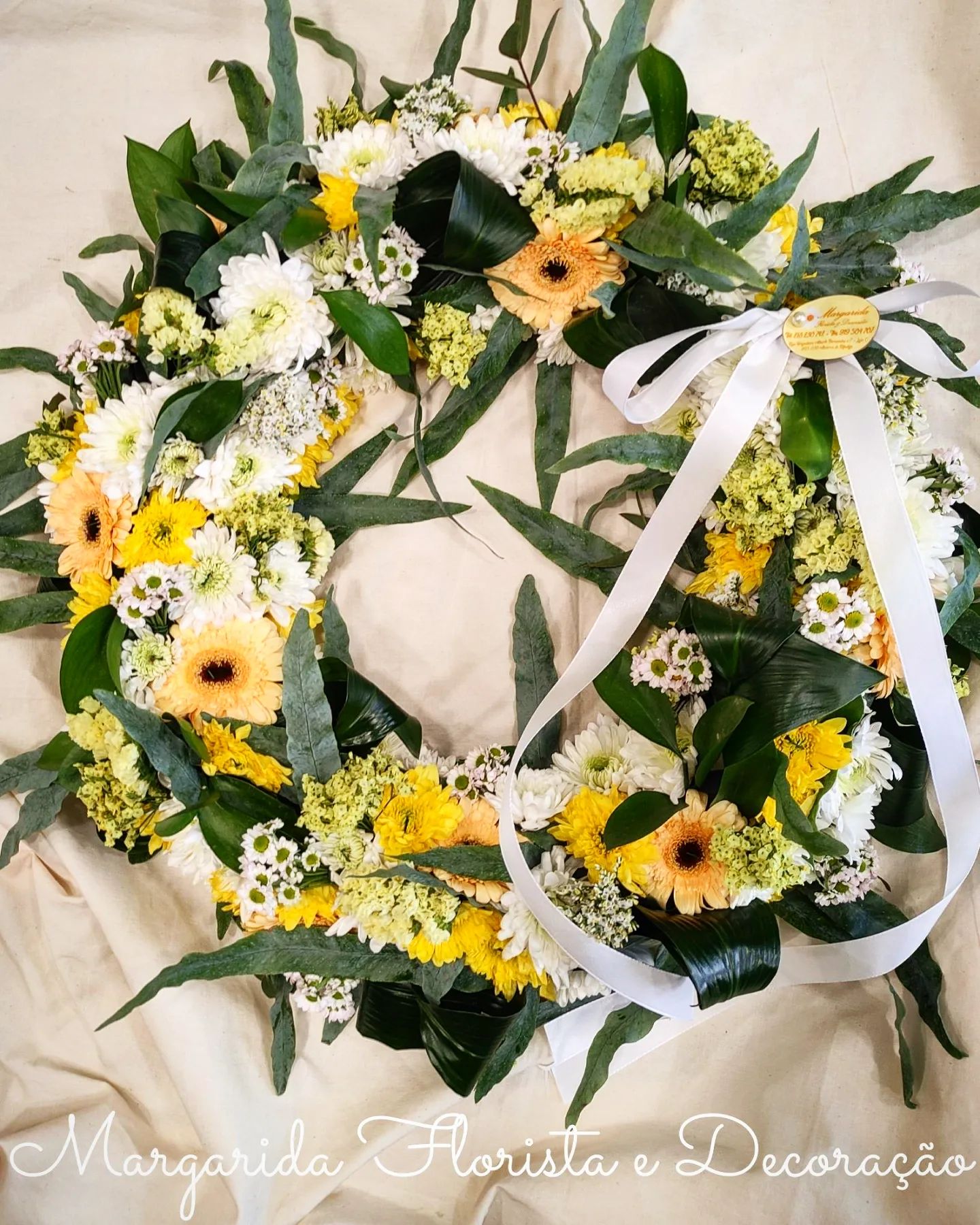 Coroa floral circular com flores amarelas e brancas e folhas verdes, decorada com fita branca