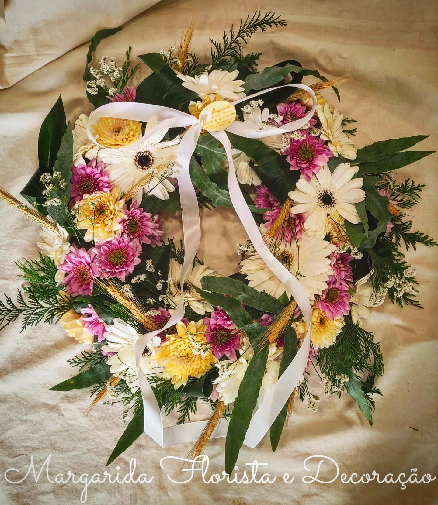 Coroa floral com flores naturais coloridas, folhas verdes e laço branco sobre tecido claro