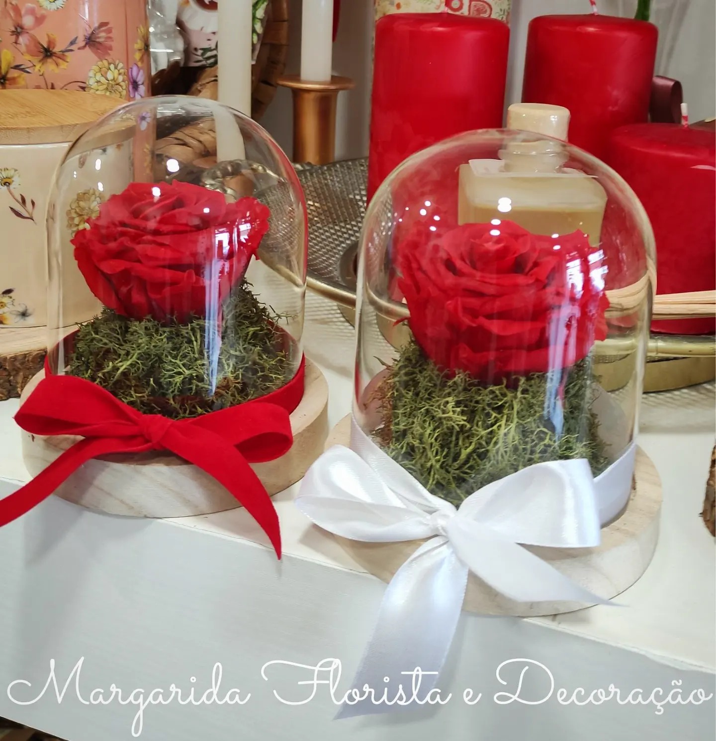 Dois arranjos florais com rosas vermelhas sob campânulas de vidro e fitas coloridas.