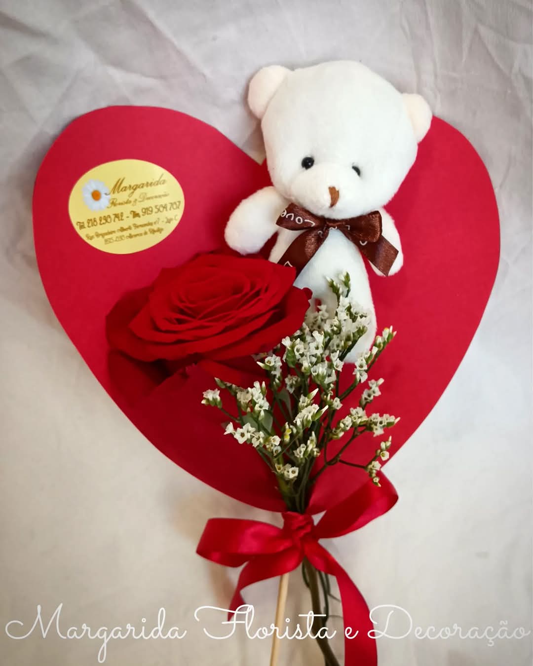 Arranjo com urso de peluche, rosa vermelha e flores brancas sobre coração vermelho