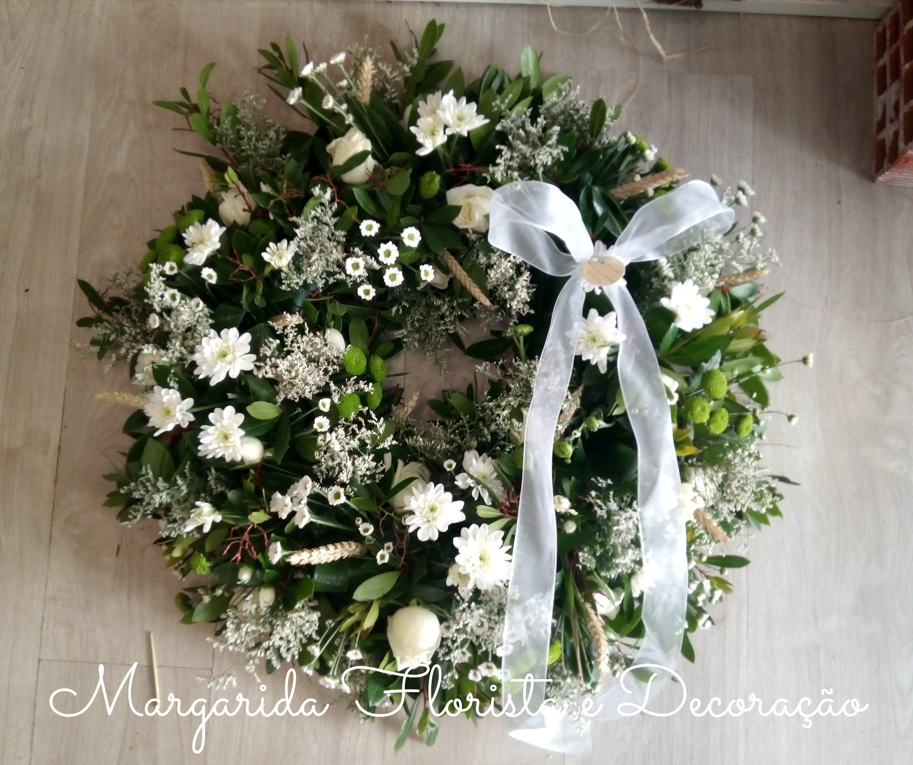 Coroa floral decorativa com folhas verdes, flores brancas e fita branca sobre chão de madeira