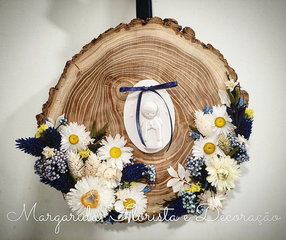 Peça decorativa circular de madeira com flores brancas, amarelas e azuis e figura oval branca pendurada ao centro