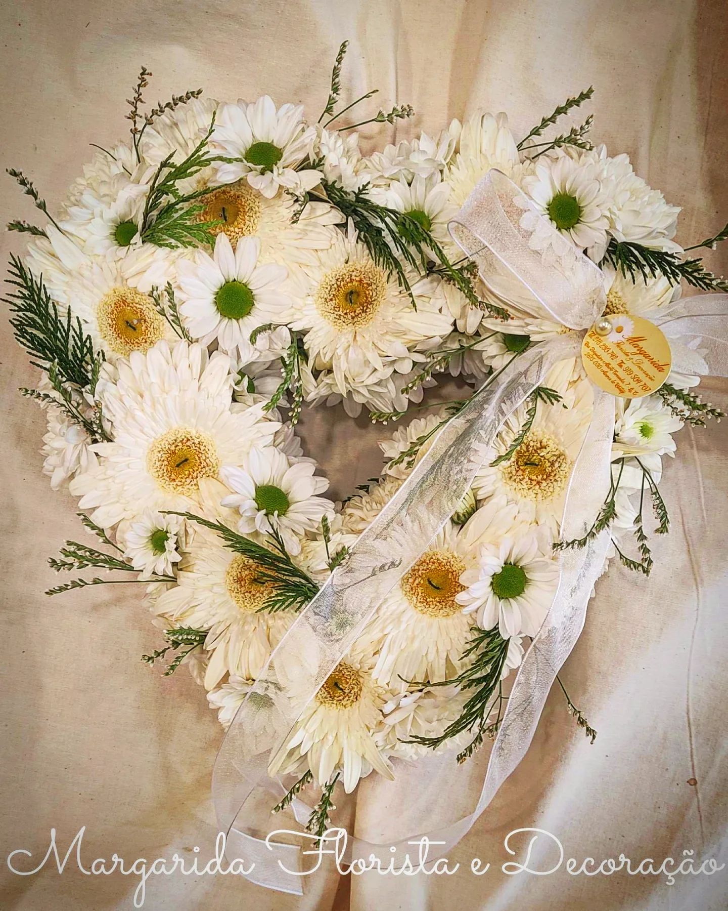 Coroa em forma de coração com margaridas brancas, laço branco e cartão amarelo sobre fundo bege