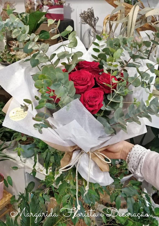 Buquê de rosas vermelhas e folhagem verde envolto em papel branco segurado pela mão de uma pessoa