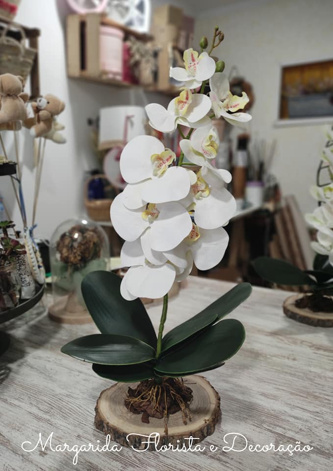 Orquídea artificial branca com folhas verdes em base de madeira sobre mesa em ambiente decorado