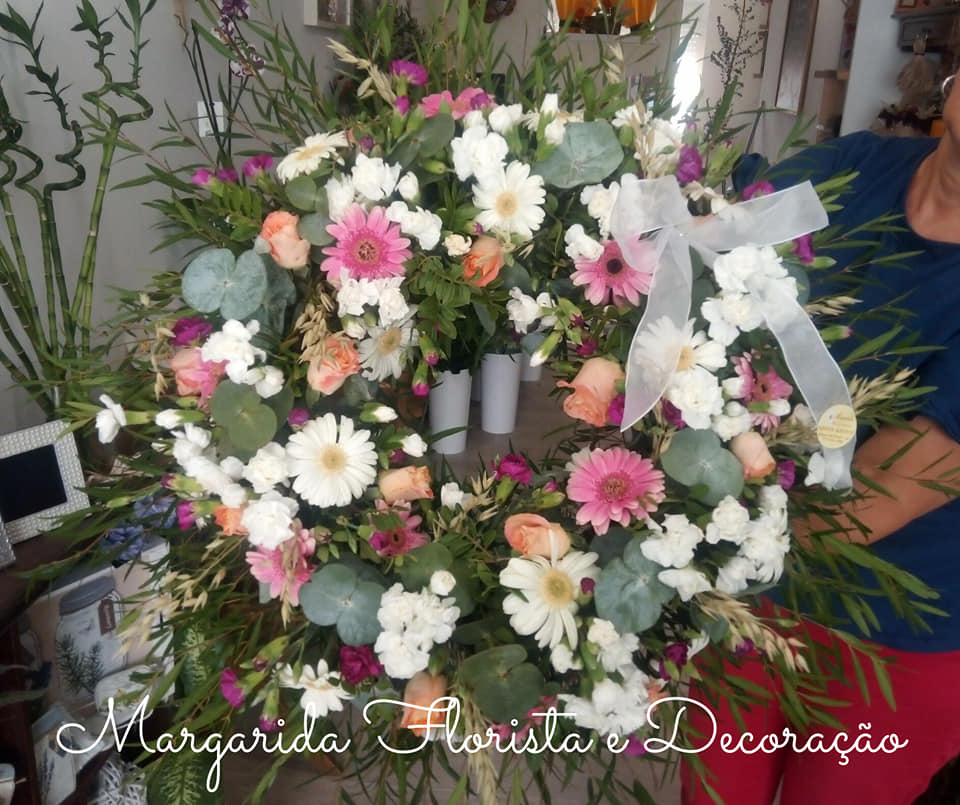 Coroa floral colorida com laço branco em ambiente interior com plantas.