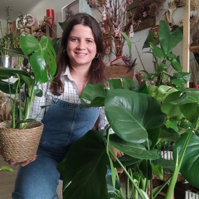Mulher sorridente sentada entre plantas verdes em vasos, interior de casa decorado com plantas e objetos naturais.