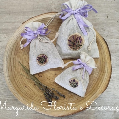 Sacos de tecido bege com laços lilás e flores, sobre prato de madeira redondo com ramo de lavanda