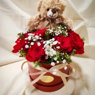 Arranjo floral com rosas vermelhas, flores brancas e ursinho de pelúcia numa caixa redonda.