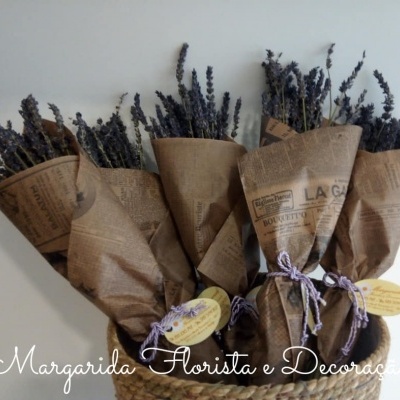 Cinco bouquets de flores secas em papel tipo jornal castanho amarrados com fita lilás e branca numa cesta de verga