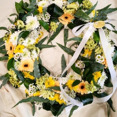Coroa floral circular com flores amarelas e brancas e folhas verdes, decorada com fita branca