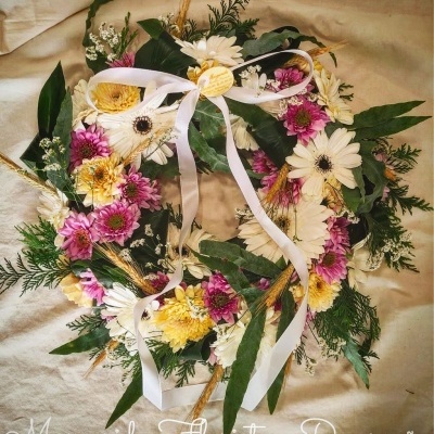 Coroa floral com flores naturais coloridas, folhas verdes e laço branco sobre tecido claro