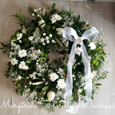 Coroa floral decorativa com folhas verdes, flores brancas e fita branca sobre chão de madeira