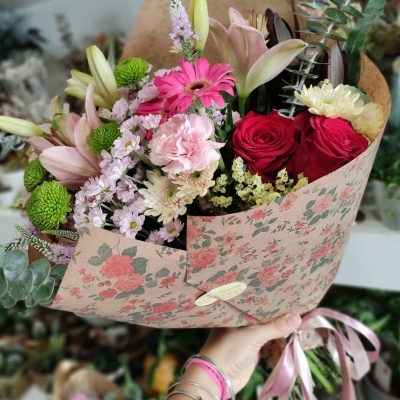 Buquê colorido de flores variadas com envelope floral rosa segurado por mão com pulseiras