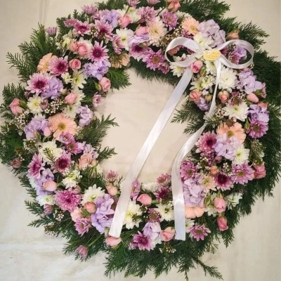 Coroa floral colorida com flores variadas e fita branca, sobre fundo claro