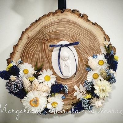 Peça decorativa circular de madeira com flores brancas, amarelas e azuis e figura oval branca pendurada ao centro