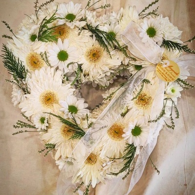 Coroa em forma de coração com margaridas brancas, laço branco e cartão amarelo sobre fundo bege