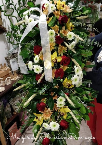 Arranjo floral vertical com rosas vermelhas, flores brancas e amarelas, folhas verdes e fita branca