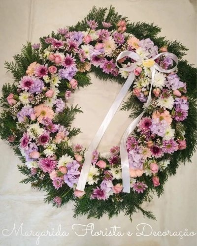 Coroa floral colorida com flores variadas e fita branca, sobre fundo claro