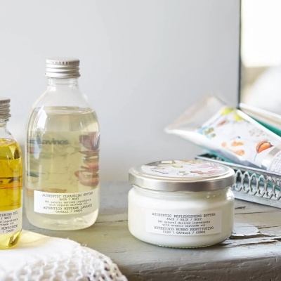 Produtos de beleza em frascos transparentes e branco com tampas prateadas sobre uma superfície cinzenta, com escova e algodão ao lado.