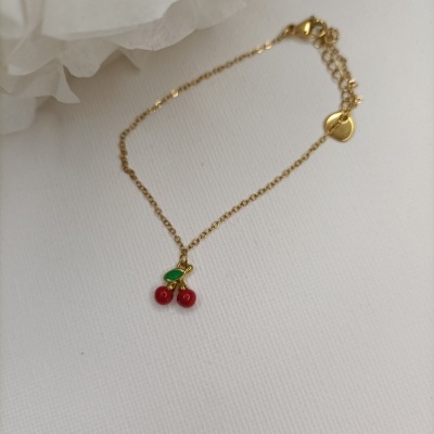 Pulseira dourada com pendente de cerejas vermelhas e folha verde sobre fundo branco