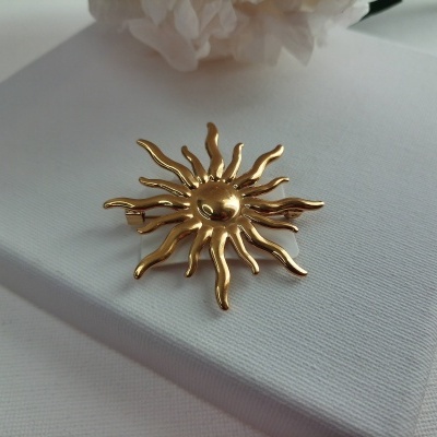 Broche dourado em forma de sol sobre superfície branca com flor branca ao fundo