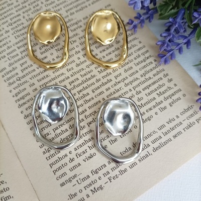 Brincos dourados e prateados com design abstrato sobre páginas de livro e flores roxas