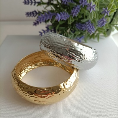 Pulseiras largas marteladas dourada e prateada com flores roxas ao fundo