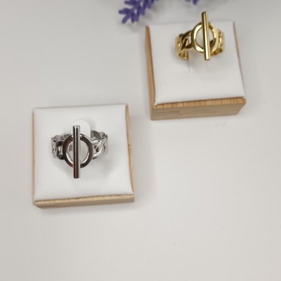 Dois anéis, prata e dourado, em exposição sobre almofadas brancas com bases de madeira, com flores roxas ao fundo.