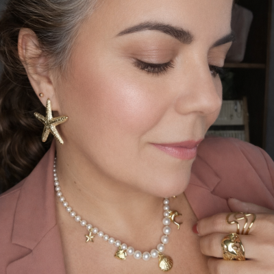 Mulher a usar brincos estrela do mar, colar de pérolas com pendentes dourados e anéis dourados