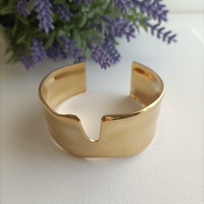 Pulseira dourada larga com abertura em V em fundo branco e flores lilás