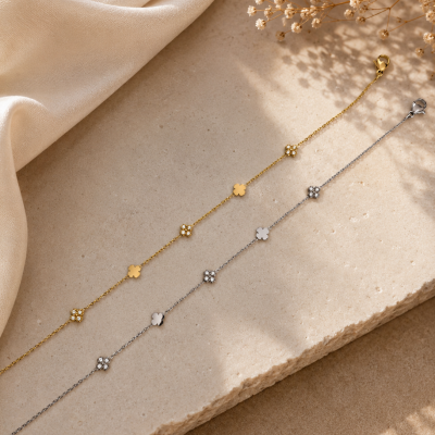 Dois colares finos dourado e prateado com pendentes em forma de flor sobre superfície de pedra clara com tecido bege e flores secas ao lado.