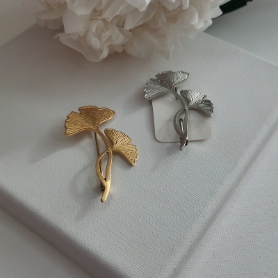 Pins decorativos em formato de folhas de ginkgo em dourado e prateado sobre superfície branca.
