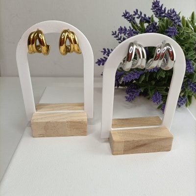 Dois pares de brincos dourados e prateados em suportes brancos com bases de madeira, com flores roxas ao fundo