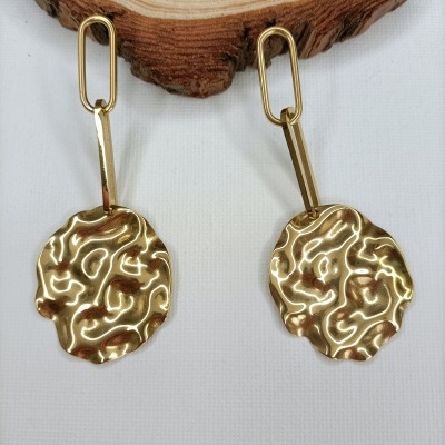 Brincos dourados ondulados com argolas retangulares sobre tecido branco e madeira clara