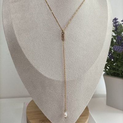 Colar dourado com pingente de pérola exposto em busto cinza claro com planta ao fundo