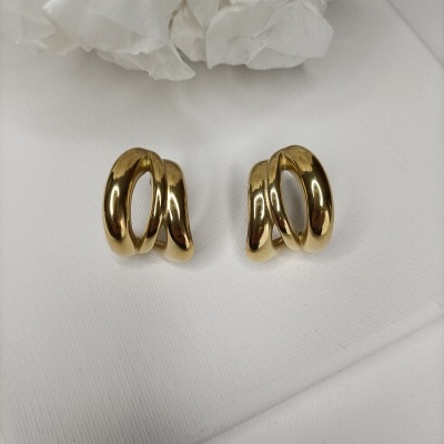 Par de brincos dourados entrelaçados sobre tecido branco com flores ao fundo