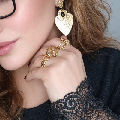 Brincos de metal dourado com textura martelada em forma de coração e anel dourado entrelaçado na mão de mulher com blusa preta de renda
