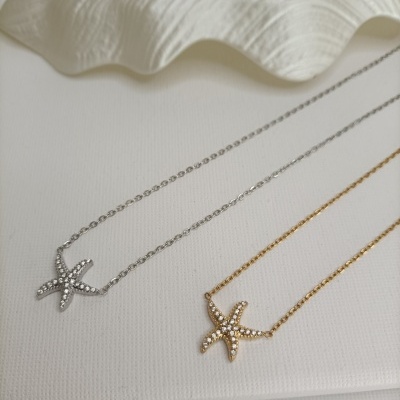 Colares com pendentes de estrela-do-mar prateado e dourado com pedras brilhantes, sobre fundo branco com concha decorativa.