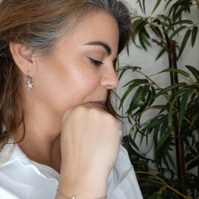 Mulher com brinco e pulseira em forma de estrela-do-mar, camisa branca e planta verde ao fundo