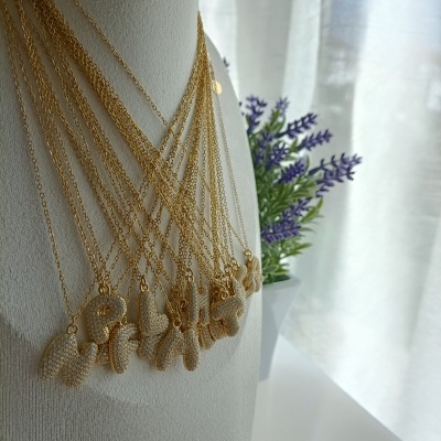 Colares dourados com letras em manequim branco, flores roxas e cortinas ao fundo