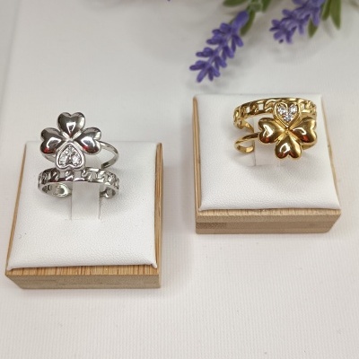 Dois anéis com trevo, um prateado e outro dourado, expostos em suportes brancos e base de madeira com flores lilases ao fundo