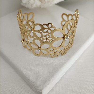 Pulseira dourada com padrão de flores e pedras transparentes sobre suporte branco.