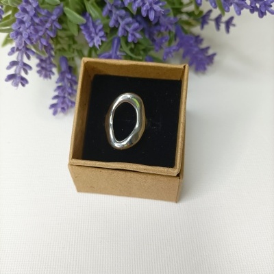 Anel prateado ovalado com caixa de cartão e flores roxas ao fundo