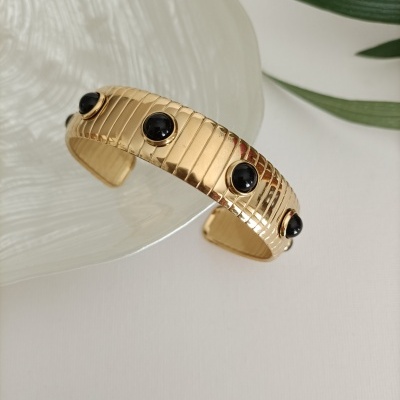 Pulseira dourada texturizada com pedras pretas redondas, sobre prato de vidro e folhas verdes ao lado.