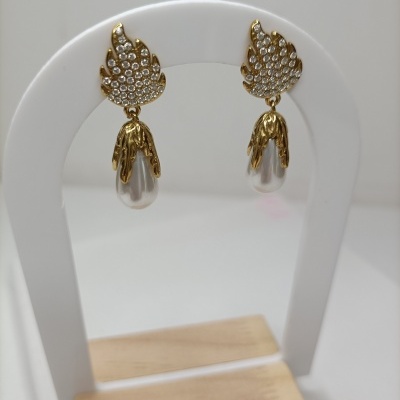 Brincos dourados com pedras e pérolas pendurados em suporte branco com base em madeira.