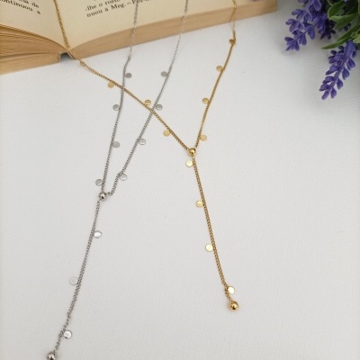 Dois colares finos dourado e prateado com discos metálicos ao longo da corrente, livro e flores lilases ao lado.