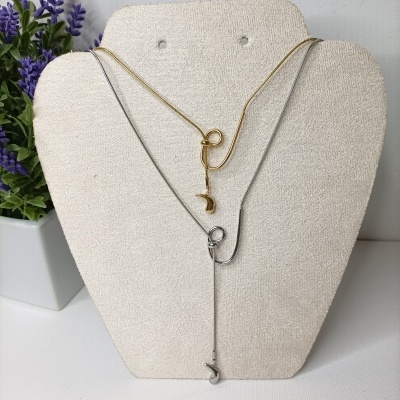 Dois colares minimalistas dourado e prateado com pingentes sobre suporte de tecido bege ao lado de vaso com planta artificial